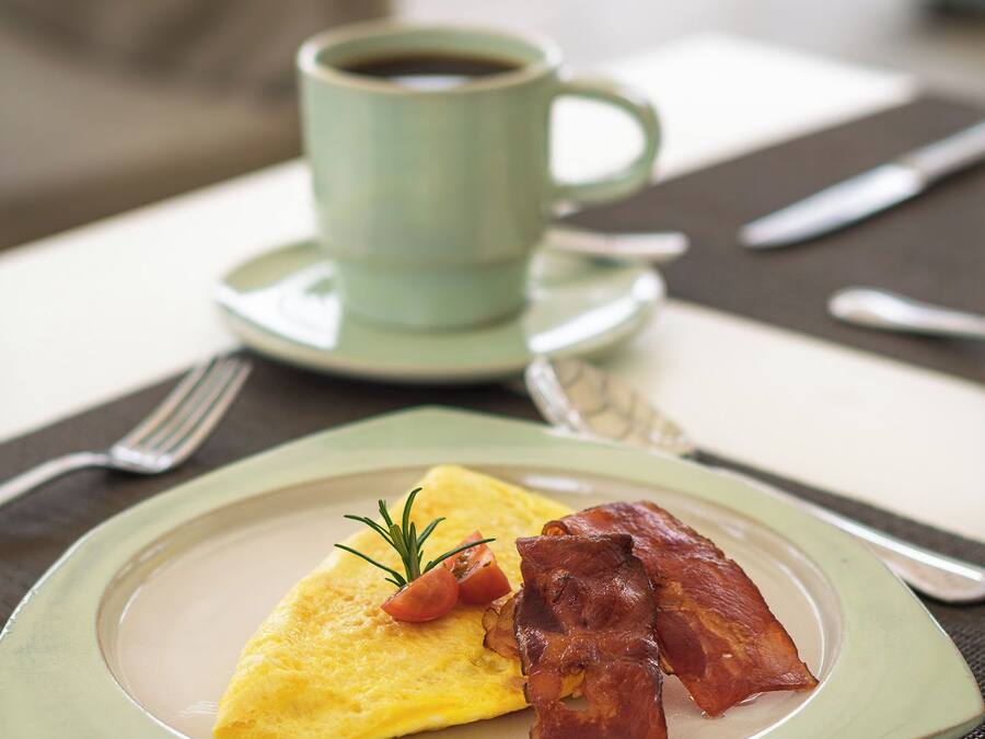 Un plato con tortilla, tocino y una ramita de romero, acompañado por una taza de café sobre una mesa.