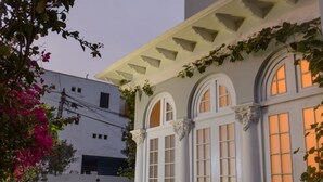 Un edificio de dos pisos con fachada blanca y ventanas arqueadas, rodeado de setos cuidados y plantas con flores.