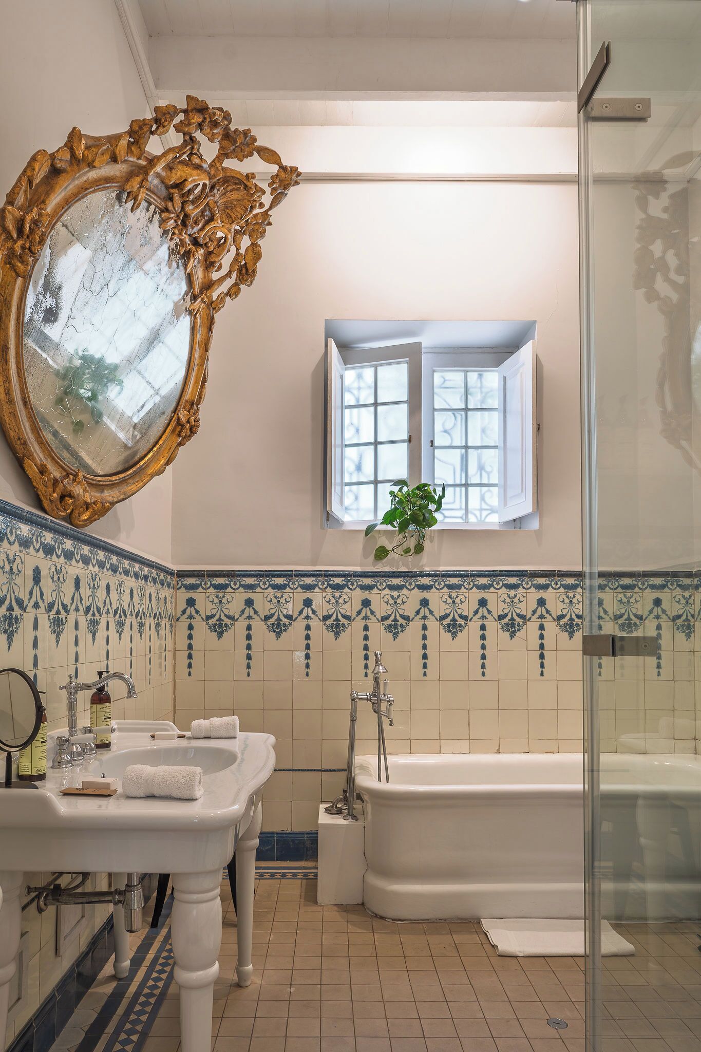 A bathroom with a large ornate mirror, a white bathtub, a sink with two towels, and a window with a plant.