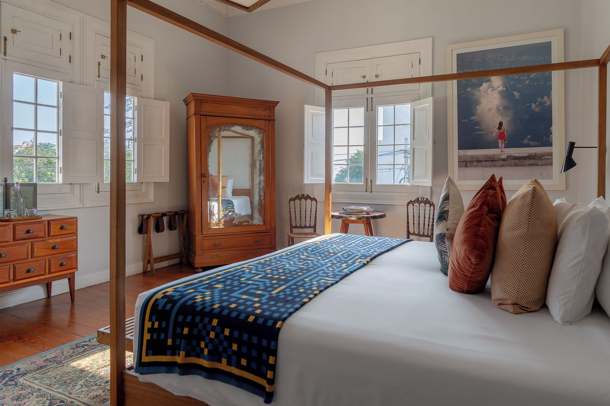 A bedroom with a large bed, a wooden wardrobe, a chair, a small table, and a painting on the wall.