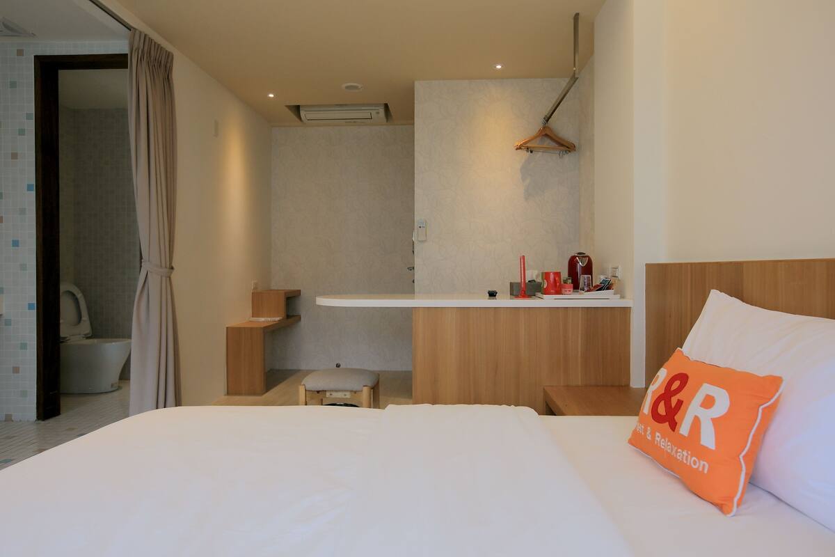 A hotel room with a bed, a bedside table, a small stool, a wooden bench, a hanging coat rack, a shower area, and a bathroom with a toilet and sink.