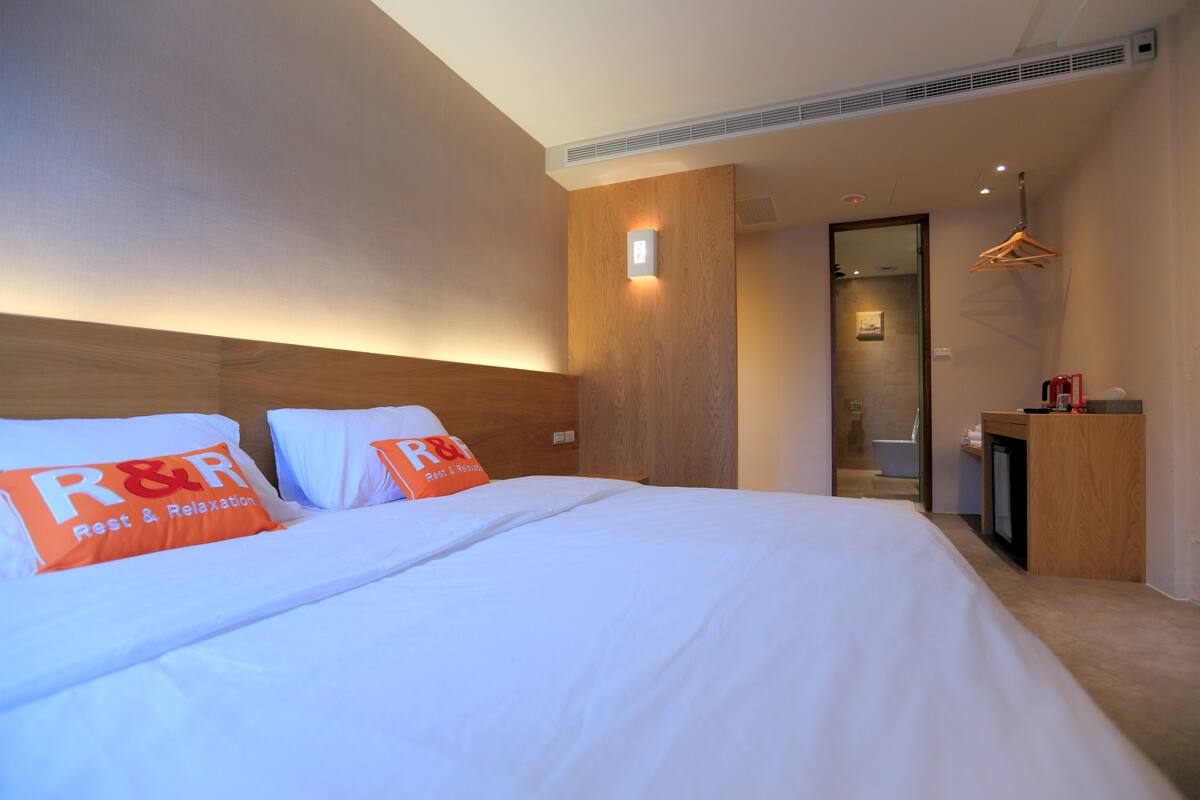 A hotel room with a large bed, two pillows with 'R&R Rest & Relaxation' branding, a wooden headboard, a bedside table, and a mirror.