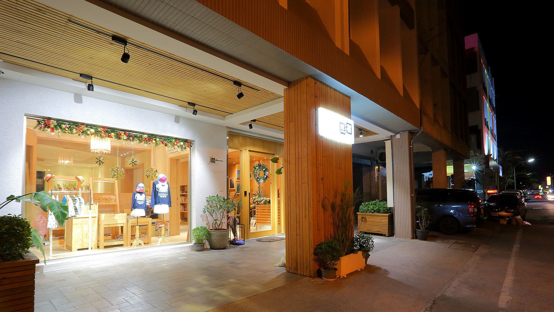 Un escaparate luminoso con fachada de madera, un letrero visible y plantas en macetas en el exterior.