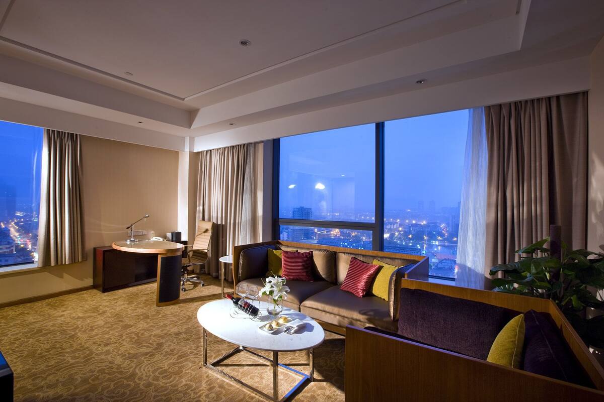 A modern hotel room with a large window, a sofa, a coffee table, and a desk with a chair.