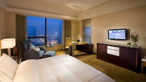 A hotel room with a large bed, a desk with a chair, a TV, and a view of the city at dusk. - Shuguang International Hotel Huaian (Huai'an)
