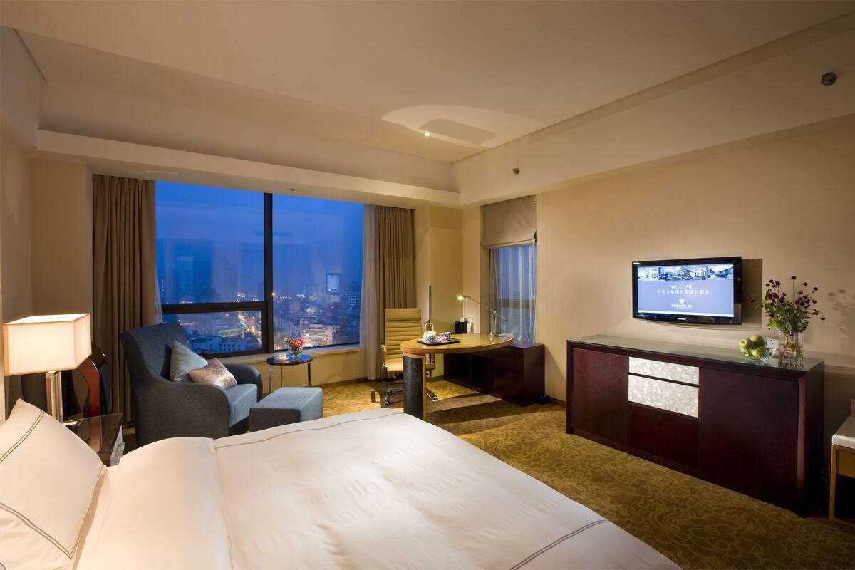 A hotel room with a large bed, a desk with a chair, a TV, and a view of the city at dusk.