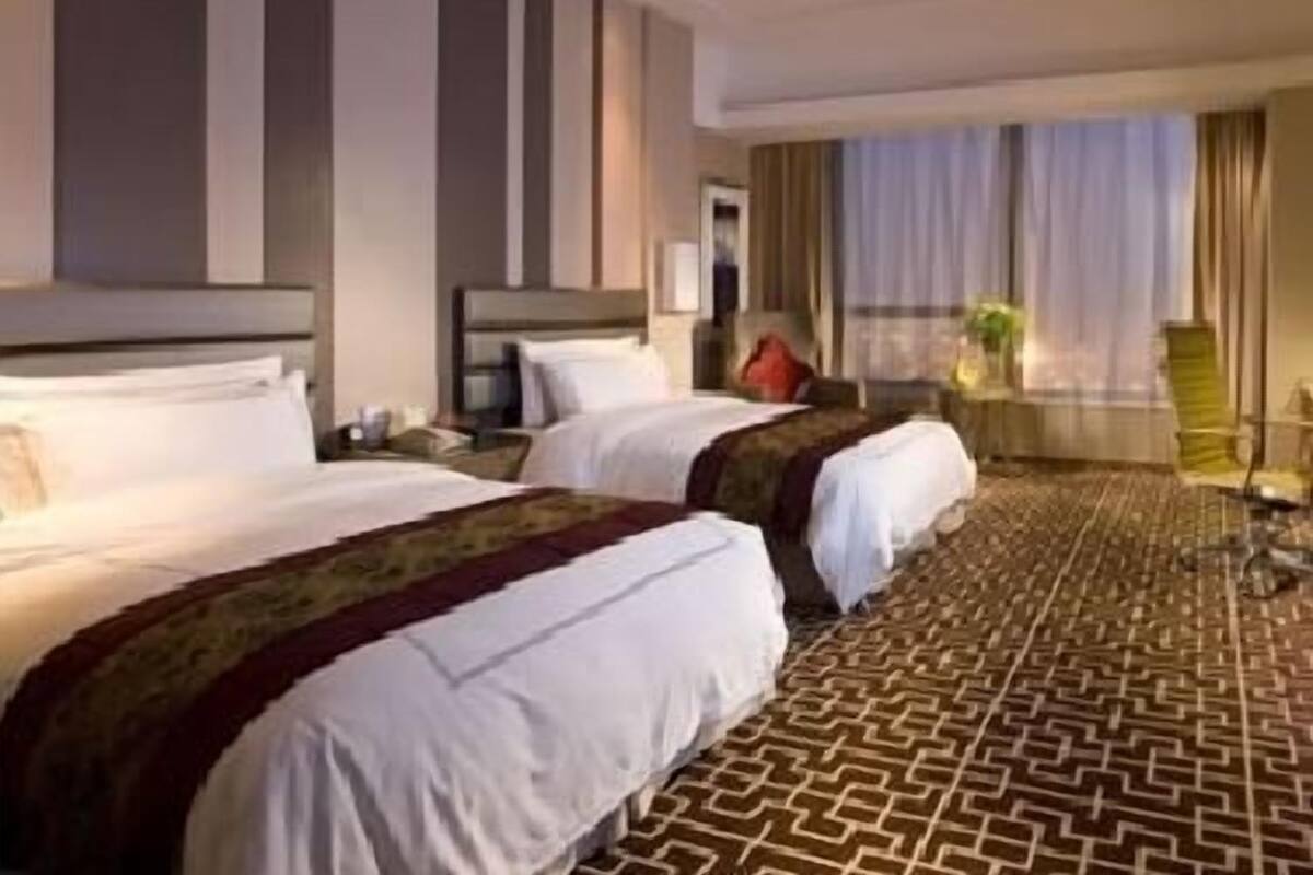 A hotel room with two beds, a large window with curtains, a patterned carpet, and a small table with a chair.