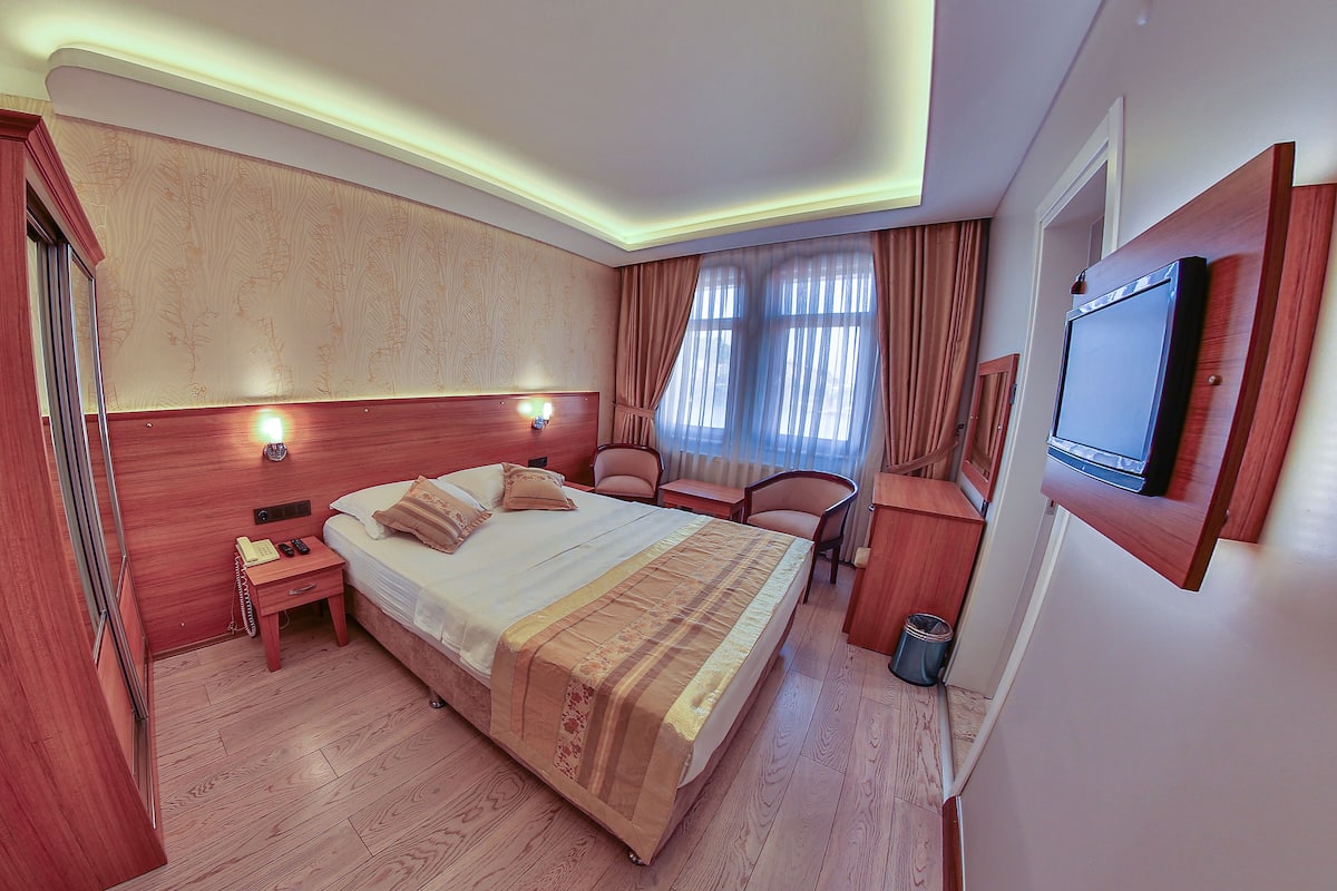A hotel room with a large bed, bedside tables, a desk, chairs, a TV, and a window with curtains.