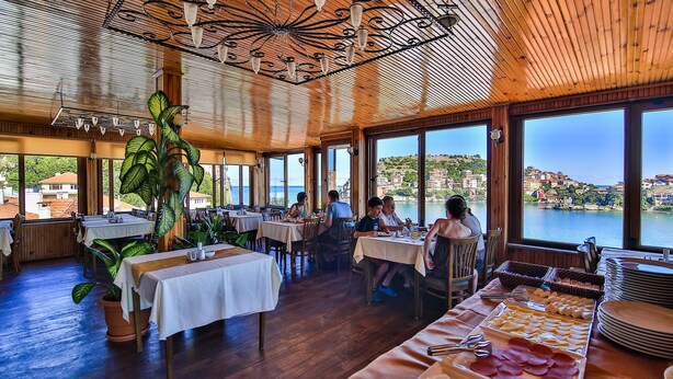 A restaurant with wooden ceilings and large windows overlooking a scenic coastal view. Tables are set with white tablecloths, and there are various food items displayed on the tables. - Seymen Hotel (Amasra)
