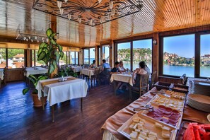 A restaurant with wooden ceilings and large windows overlooking a scenic coastal view. Tables are set with white tablecloths, and there are various food items displayed on the tables. - Seymen Hotel (Amasra)