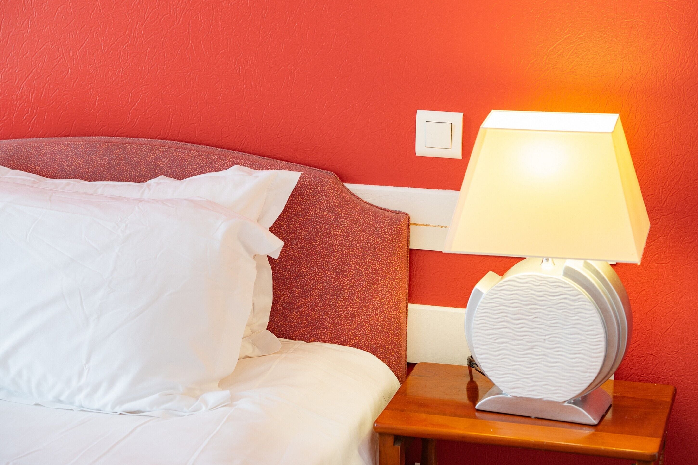 Una cama bien hecha con sábanas blancas, un cabecero rojo con textura y una mesita de noche con una lámpara.