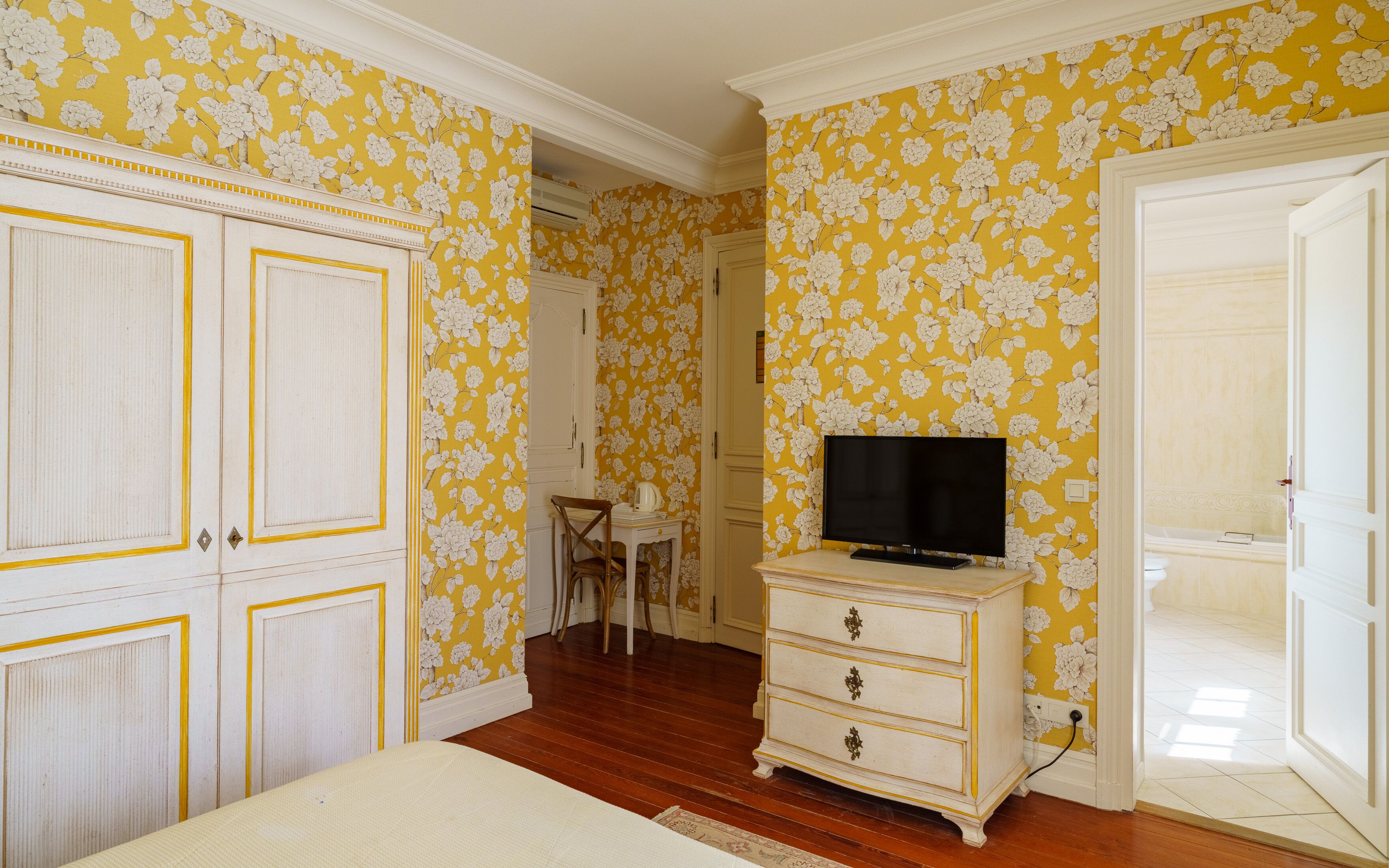 A bedroom with floral wallpaper, a wooden dresser, a television, and a wardrobe.