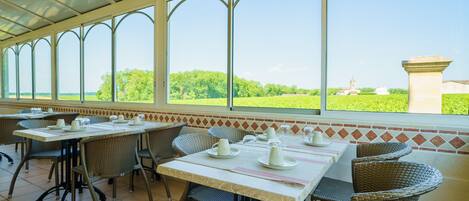 A covered outdoor dining area with tables and wicker chairs, large windows offering a view of greenery and a distant building.