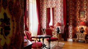 Een kamer met decoratief behang, rode gordijnen en klassiek meubilair, waaronder een tafel en stoelen.