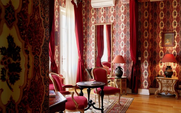 A room with ornate wallpaper, red curtains, and classic furniture including a table and chairs.