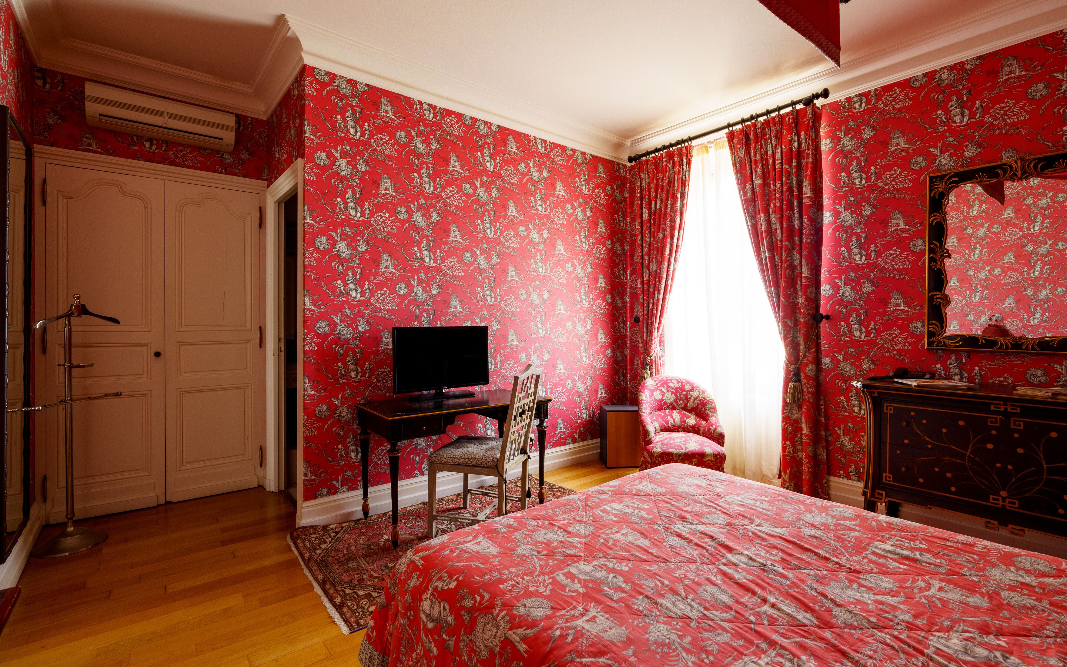A bedroom with a large bed, a desk with a chair, a television, and a wardrobe.