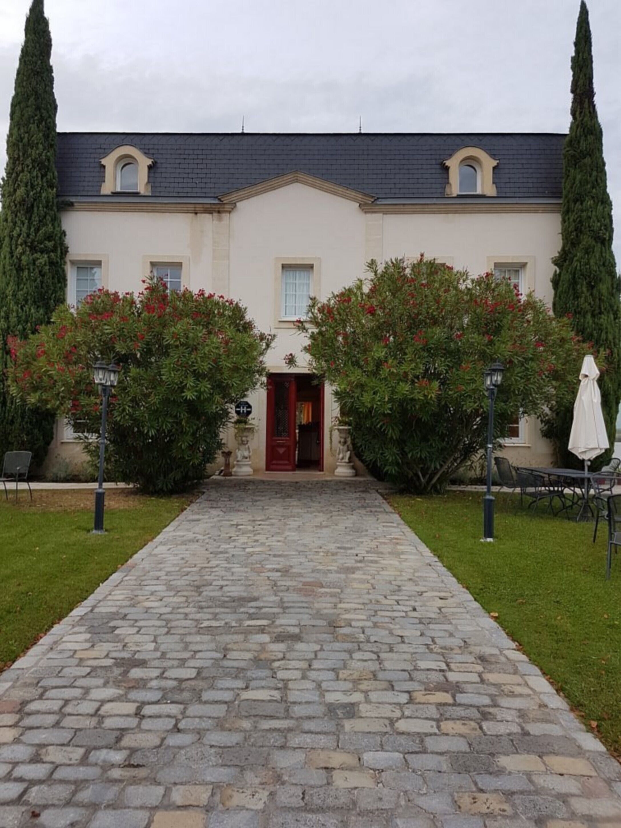 A building with a central red door, flanked by two trees with red flowers, and a cobblestone pathway leading to the entrance.
