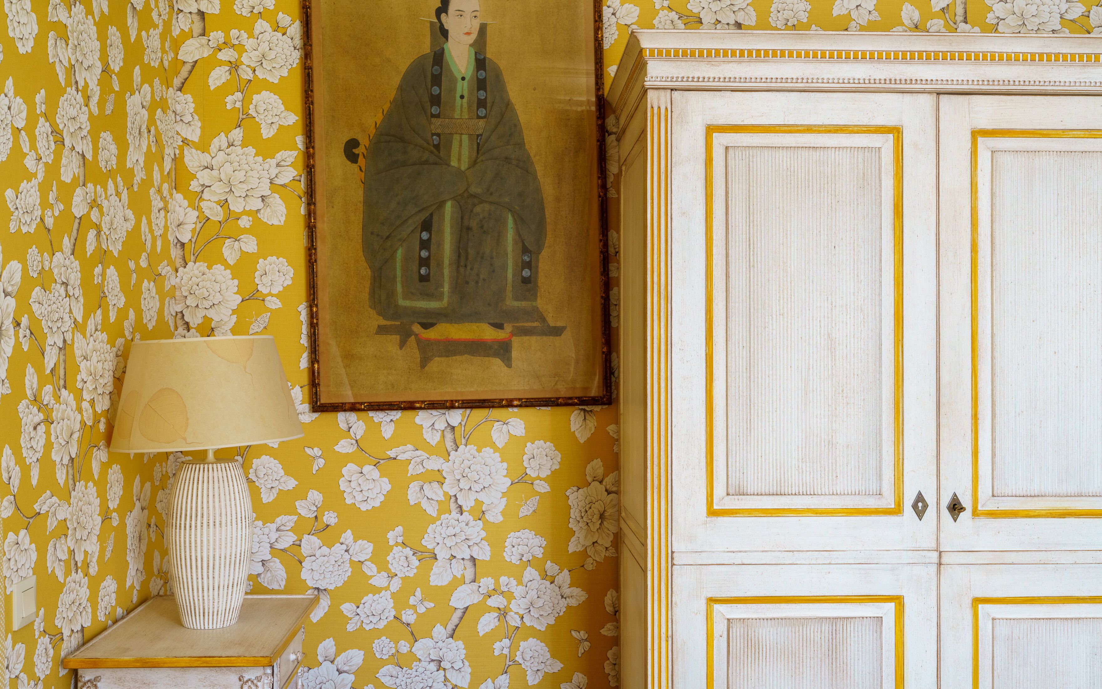 A room with yellow floral wallpaper, a framed painting, a lamp, and a white cabinet with gold trim.