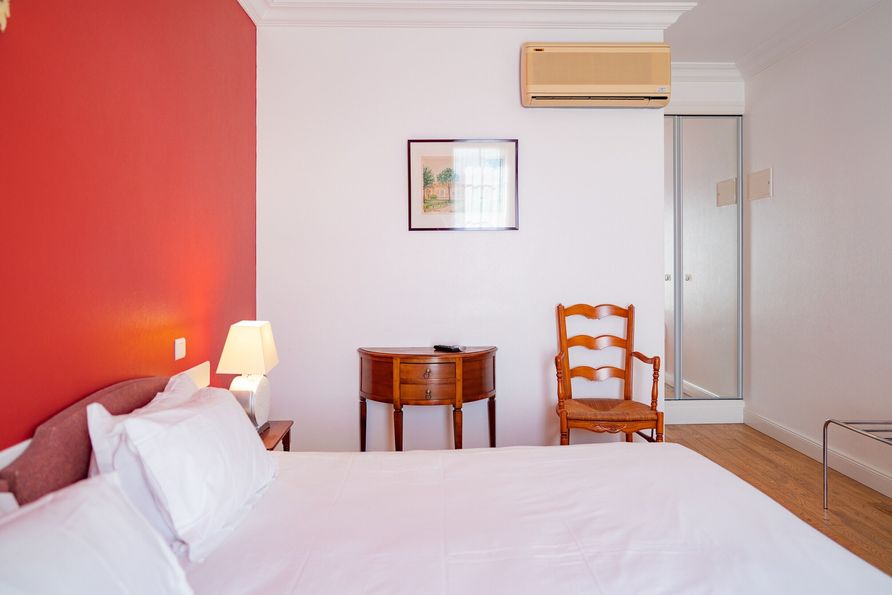 Una habitación de hotel con una cama, una silla, una mesita, una lámpara, un cuadro enmarcado y un equipo de aire acondicionado.