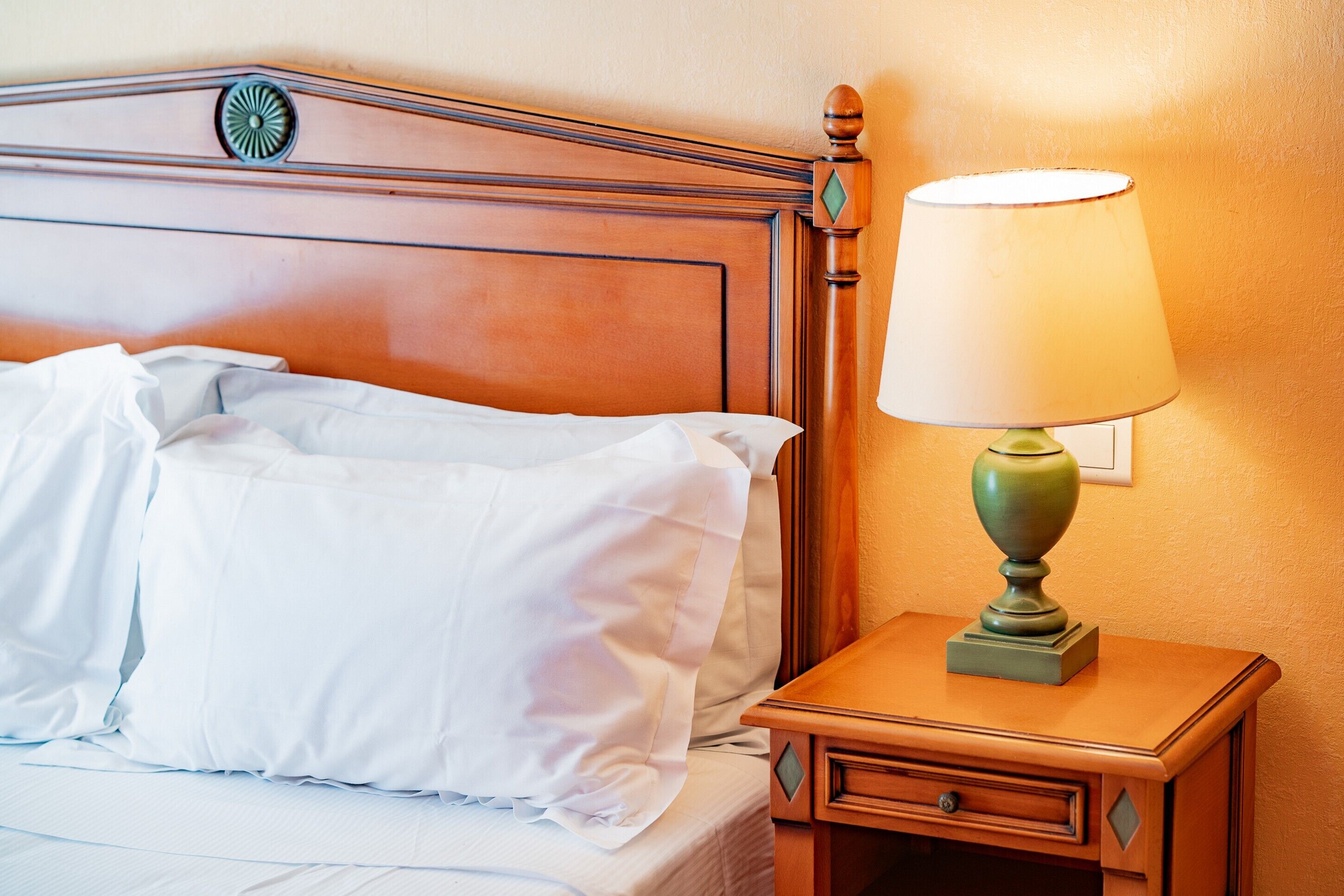 Een net opgemaakt bed met witte lakens en kussens, een houten nachtkastje met een groene lamp en een aan de muur bevestigd fotolijstje.