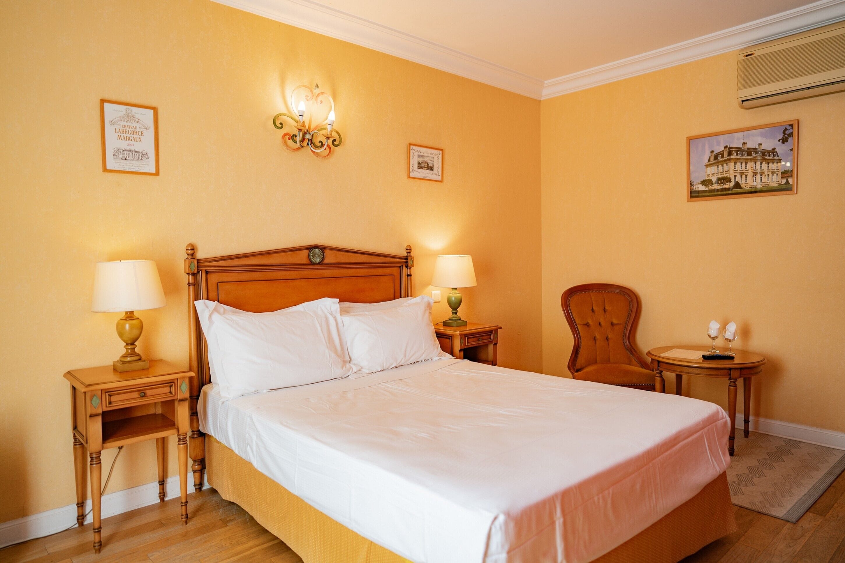 A hotel room with a large bed, two bedside tables with lamps, a chair, and a small table with two wine glasses.