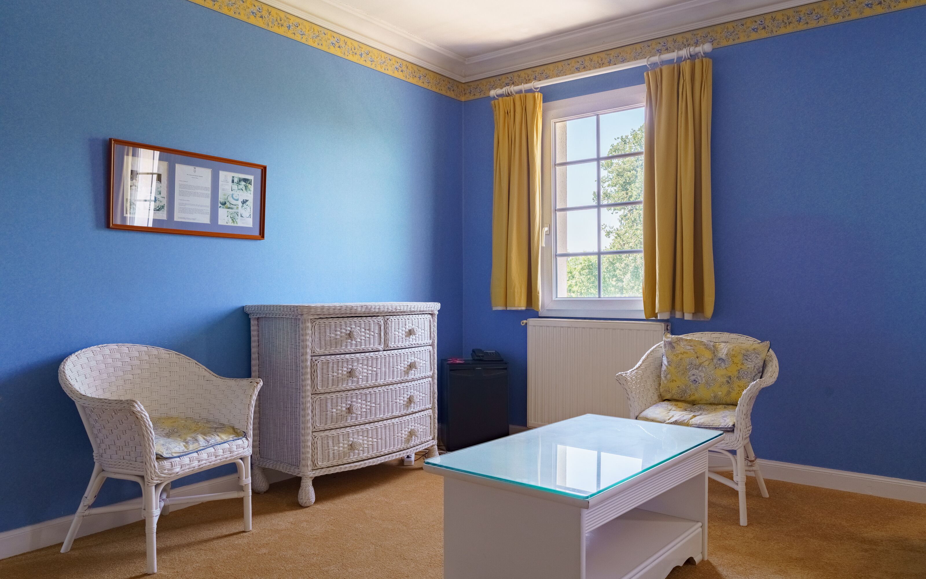 Una habitación con paredes azules, un aparador blanco, dos sillas de mimbre, una mesa con superficie de vidrio y una ventana con cortinas amarillas.