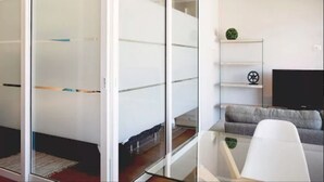 Ein modernes Wohnzimmer mit Glastrennwand, einem grauen Sofa, einem weißen Sessel, einem kleinen Tisch und einem Fernseher auf einem Standfuß.