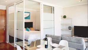 Ein modernes Hotelzimmer mit einer Glastrennwand, die Schlafbereich von Wohnbereich abtrennt. Ein Bett mit Kissen, eine Couch, ein Fernseher und ein kleiner Tisch mit Flasche und Gläsern sind vorhanden.