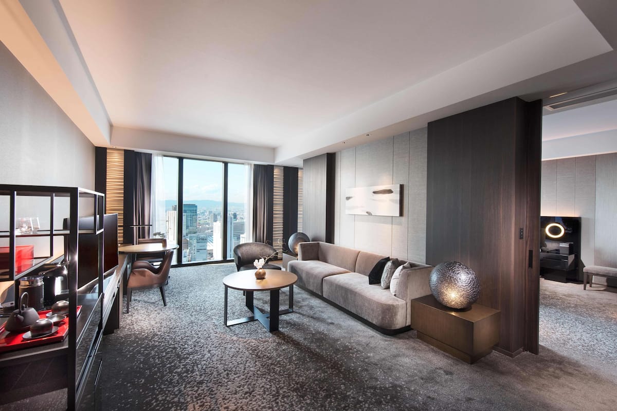 A modern hotel room with a large window, a sofa, a coffee table, and a dining area with a red table runner.