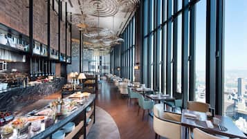 A modern restaurant with a long counter, high ceilings, and large windows offering city views.
