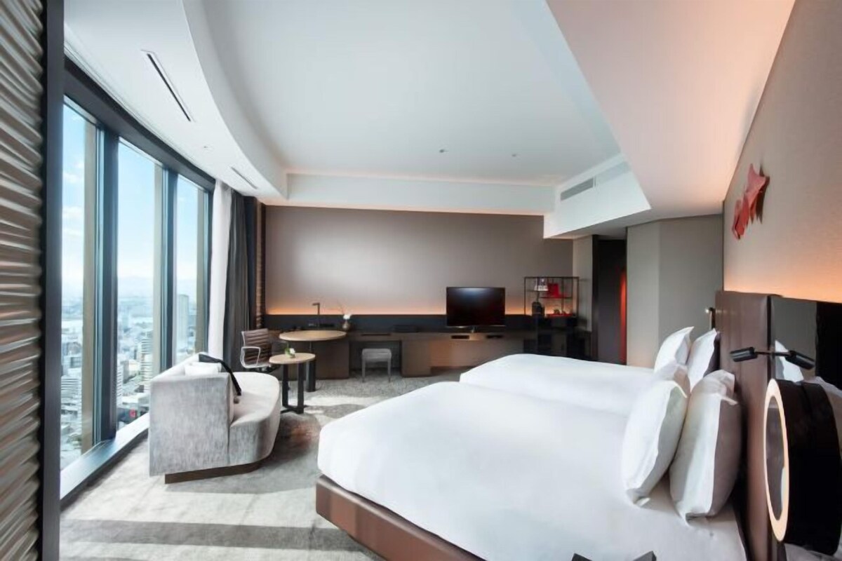 A modern hotel room with a large bed, a desk, a television, and a view of the city through large windows.
