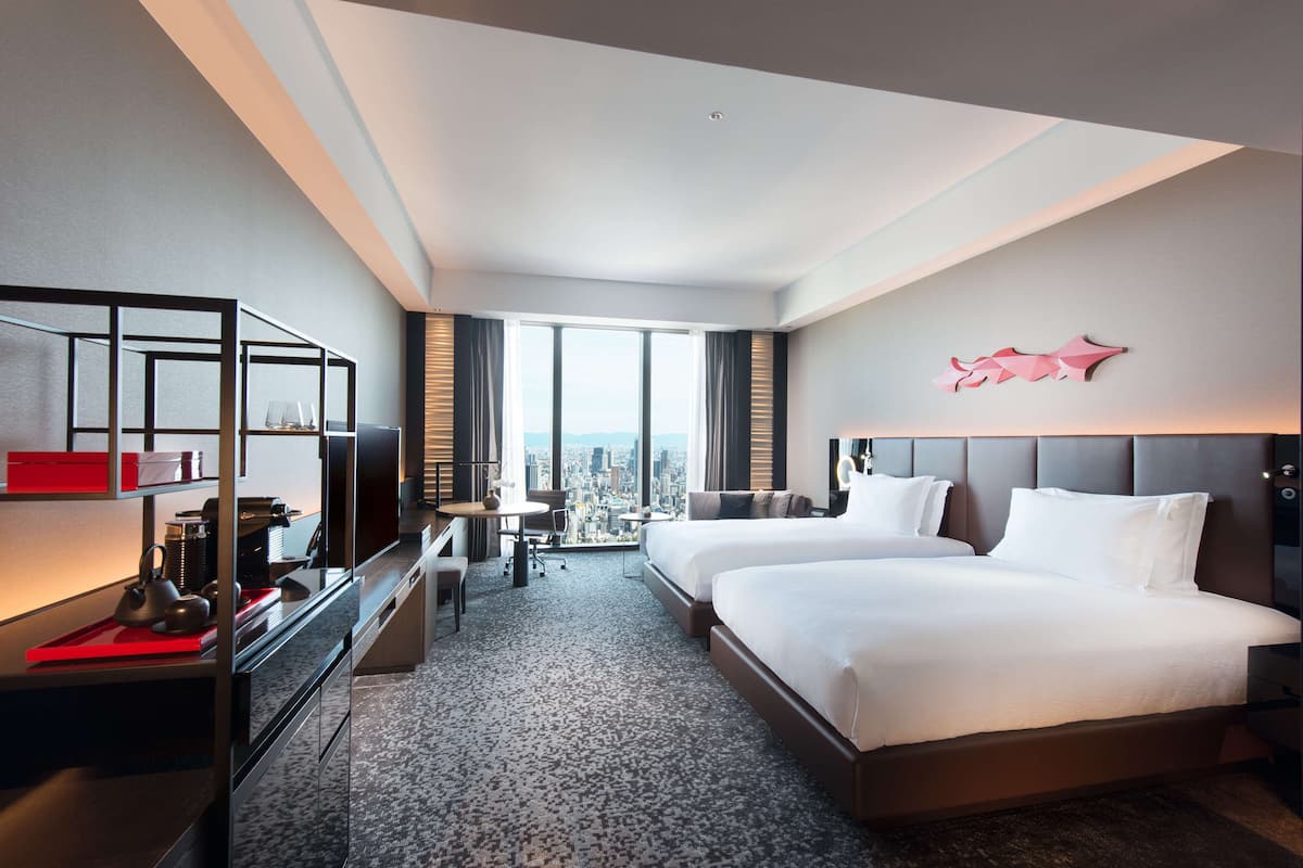 A hotel room with two beds, a desk, a chair, and a TV. There is a large window with a city view.