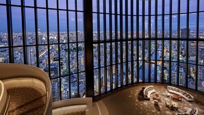 Une chambre d’hôtel située dans un immeuble de grande hauteur, offrant une vue panoramique sur la ville grâce à de grandes fenêtres du sol au plafond, un espace salon aux lignes courbes et un décor mural géométrique original.