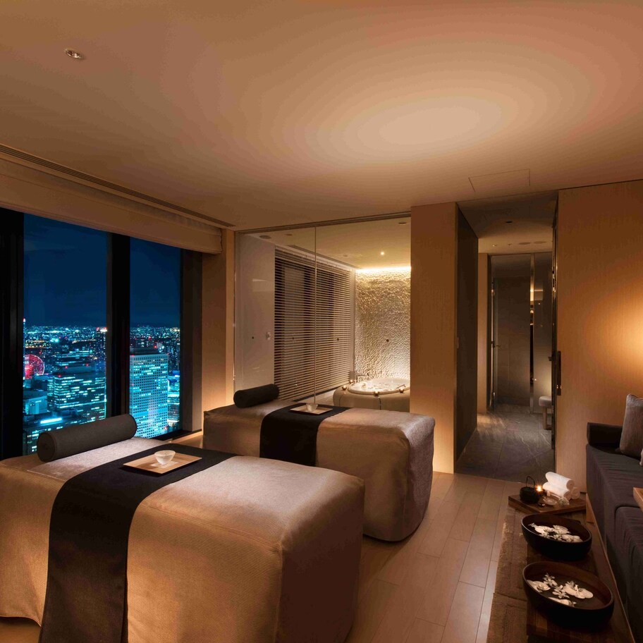 A modern hotel room with a city view, a sofa, two ottomans, and a small table with candles.