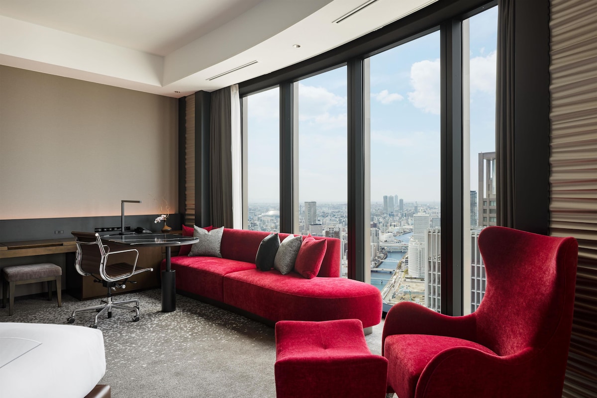 A modern hotel room with a red sofa, a desk, and a view of the cityscape.
