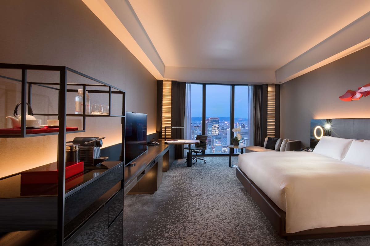 A hotel room with a bed, desk, chair, TV, and a view of the cityscape.