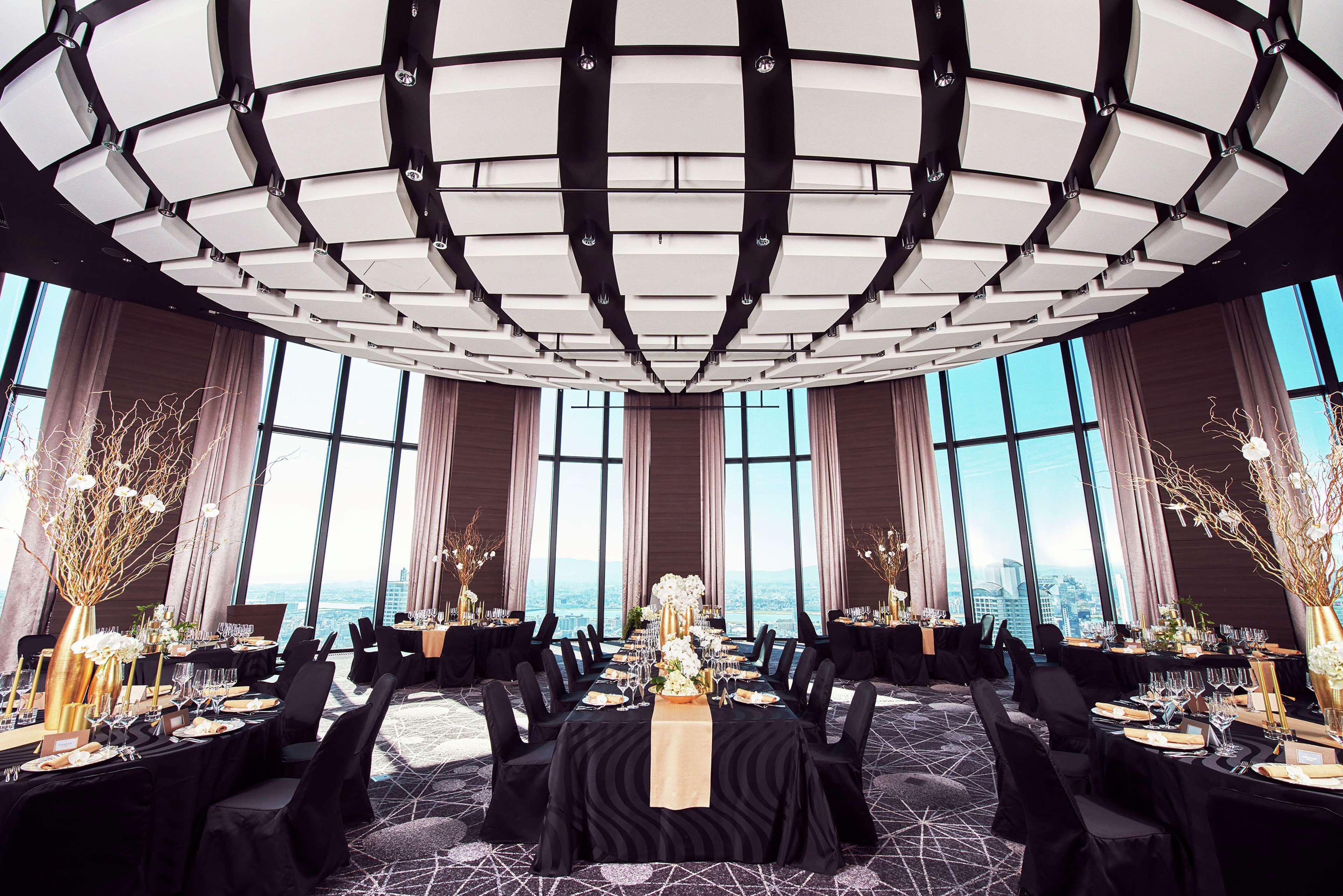 A spacious, elegantly decorated banquet hall with a high ceiling and large windows offering a city view.