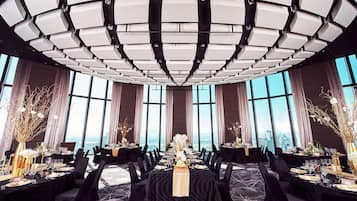 A spacious, elegantly decorated banquet hall with a high ceiling and large windows offering a city view.