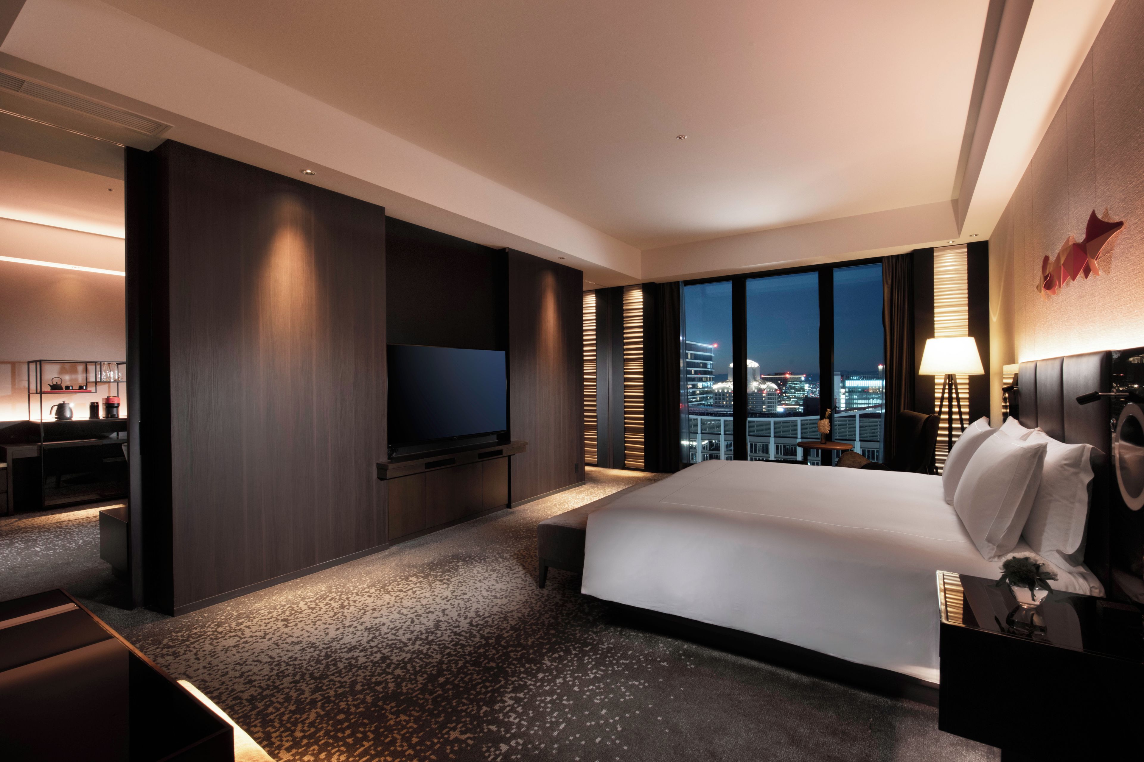 A modern hotel room with a large bed, a flat-screen TV mounted on the wall, a desk with a chair, and a view of the cityscape through large windows.