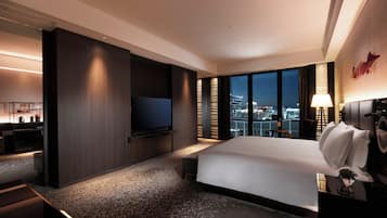A modern hotel room with a large bed, a flat-screen TV mounted on the wall, a desk with a chair, and a view of the cityscape through large windows.