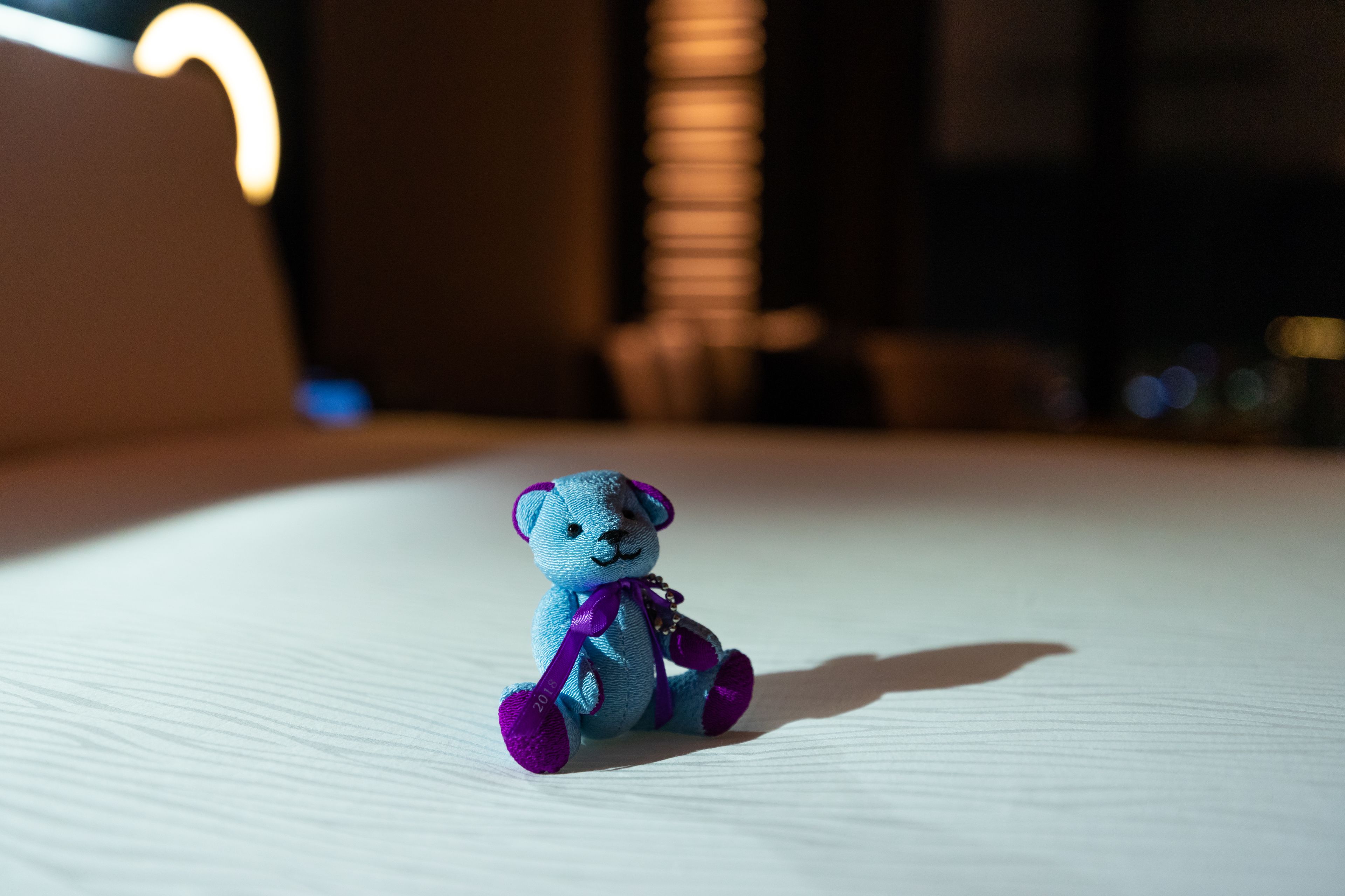 A small blue teddy bear with a purple ribbon on a white surface.