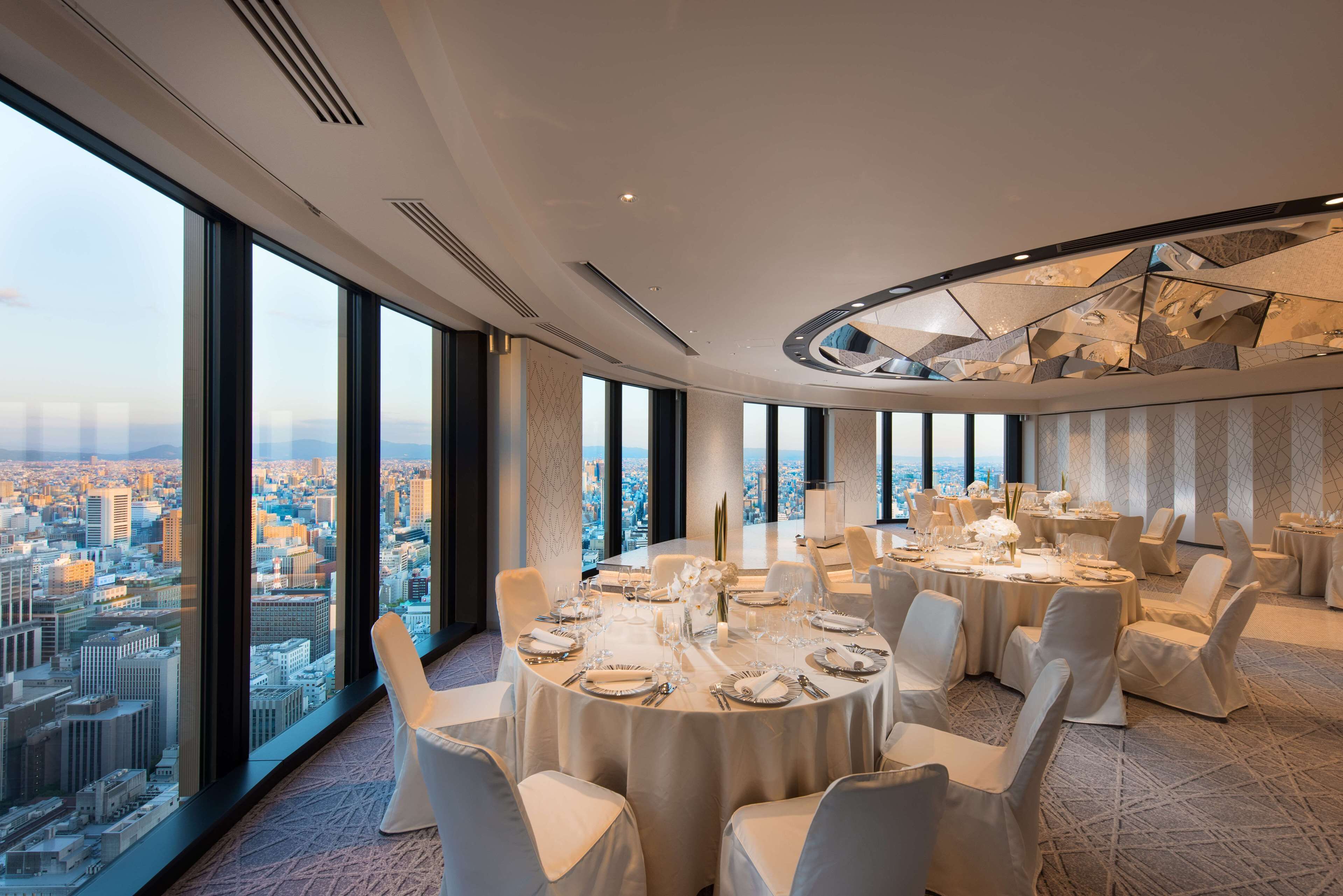 A high-rise hotel room with a panoramic city view, a round dining table set for six, and a modern ceiling design.