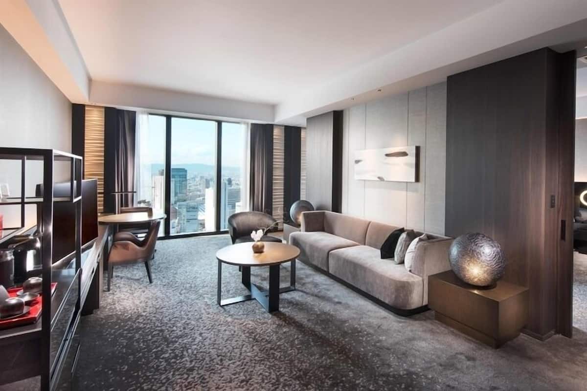 A modern hotel room with a city view, a sofa, a dining area, and a TV.