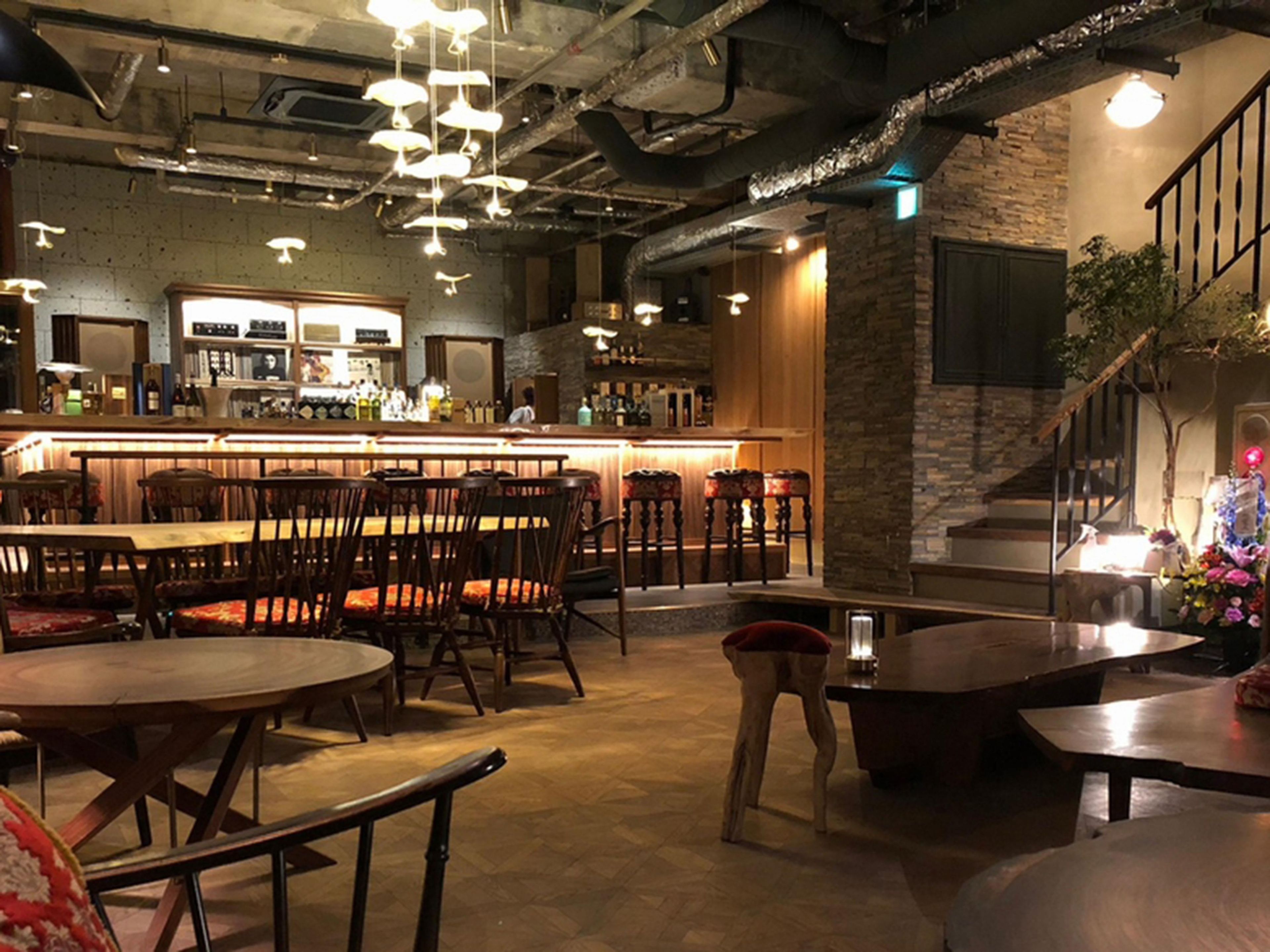 A bar area with wooden tables and chairs, a counter with bottles, and a staircase with a plant.