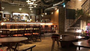 A bar area with wooden tables and chairs, a counter with bottles, and a staircase with a plant.
