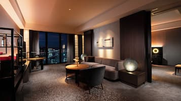 A modern hotel room with a city view, a sofa, a coffee table, and a desk with a lamp.
