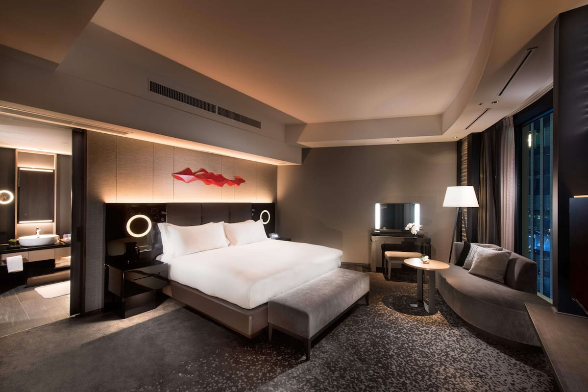 A modern hotel room with a large bed, bedside tables, a desk, a sofa, and a large window.