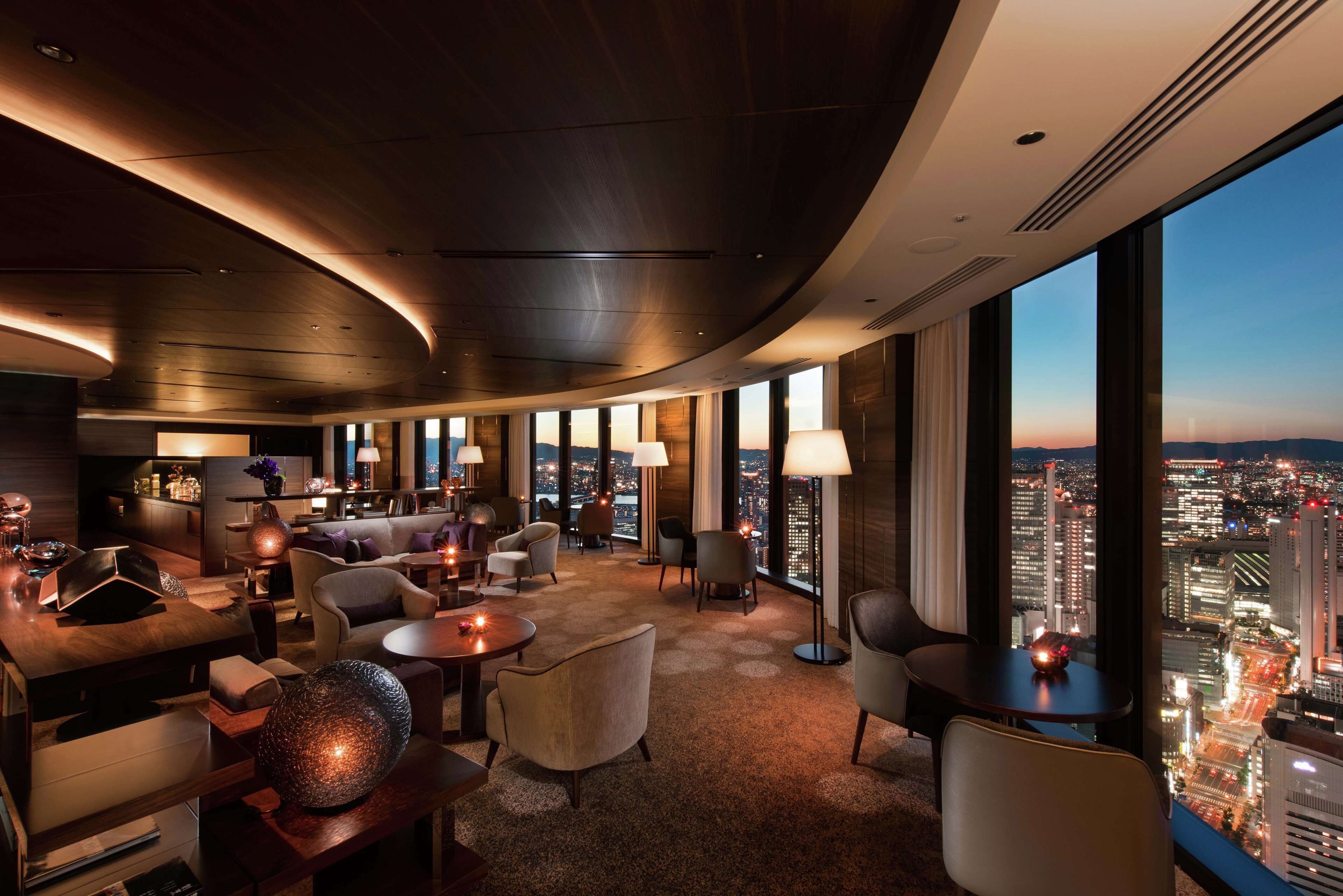 A modern hotel lobby with a curved ceiling, comfortable seating, a bar area, and a city view through large windows.