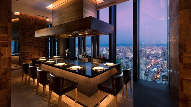 Een moderne keuken met een groot eiland, barstoelen en een stadszicht door grote ramen.
