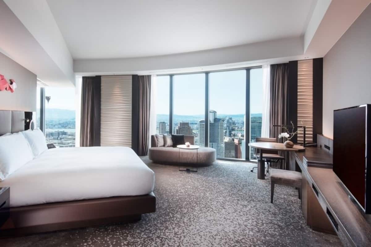 A modern hotel room with a large bed, a desk, a chair, and a TV. The room has a city view.